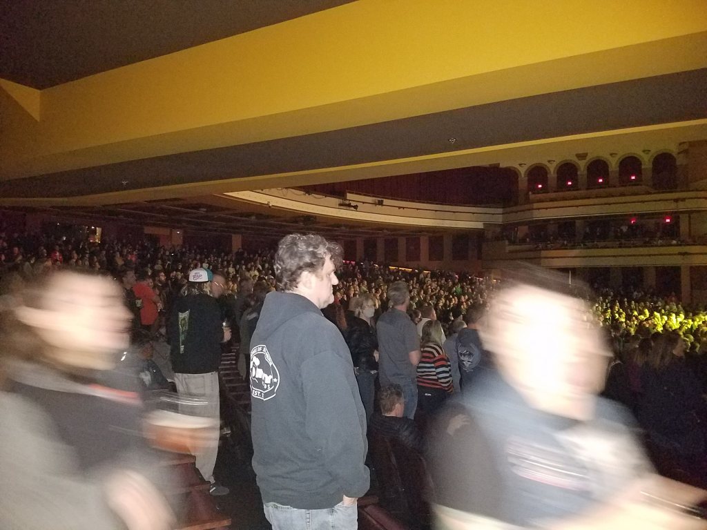 An interior shot of a theater with a crowd of people, standing, focus on a man in a sweatshirt watching the stage, which is not visible in the shot.